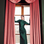 Woman in a green dress standing behind pink curtains in a room with a window.